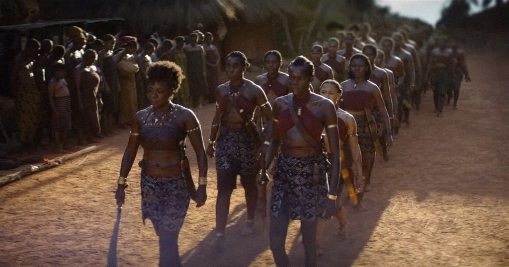 Colonne de femmes Agojiés partant en guerre : elles sont armées de machettes et portent un haut et une jupe de couleurs vives aux motifs africains.
La colonne est menée par Viola Davis. 